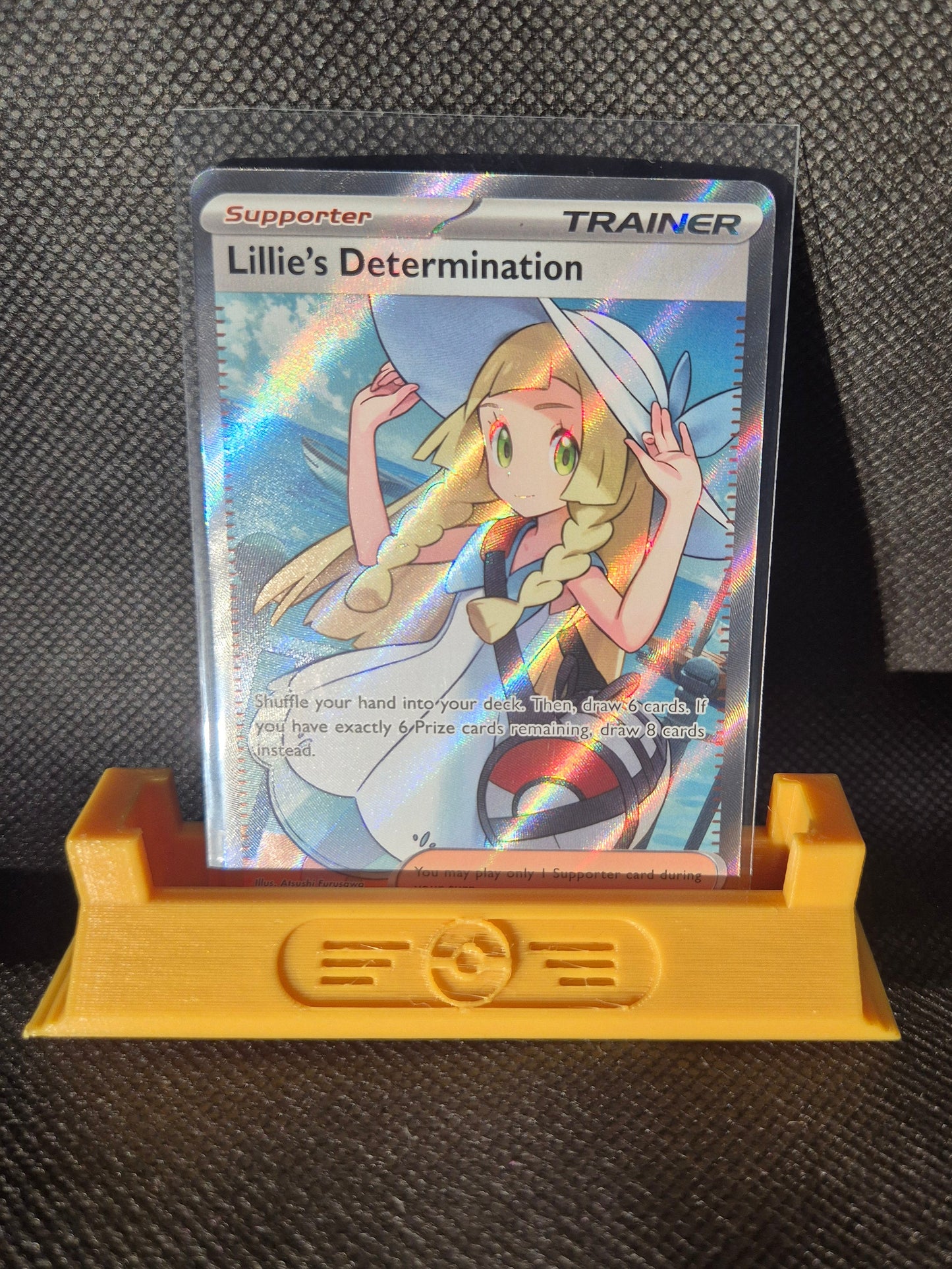 Trading card featuring a character with a hat and rainbow elements, held by a yellow card holder on a textured black surface.